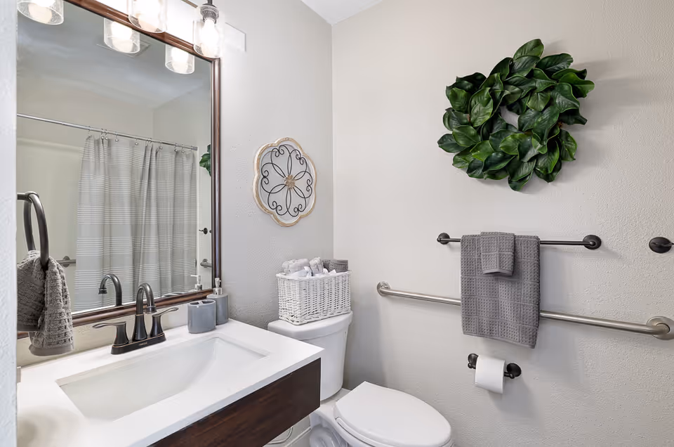 A clean and modern bathroom featuring a white sink with dark wood vanity, a large mirror with three light fixtures above, a toilet with a white wicker basket holding rolled towels on top, a gray towel hanging on a bar, a toilet paper holder, a decorative wall clock, and a green leafy wall decoration.