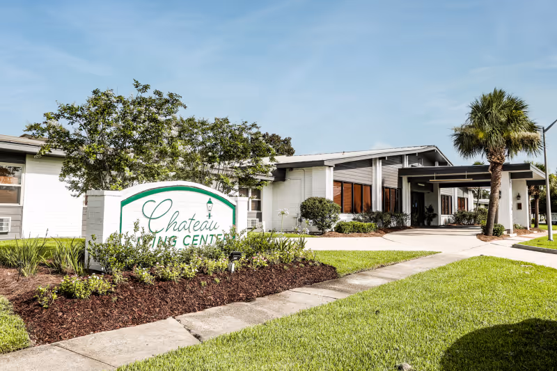 Exterior view of Chateau Living Center, showing a white building with large windows, a covered entrance, palm trees, and a landscaped garden with a sign displaying the facility's name.