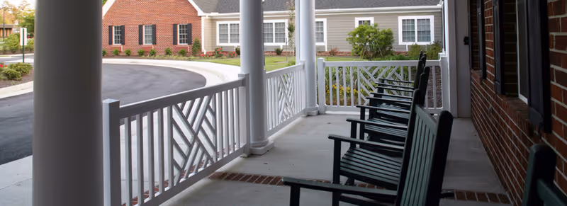 Covered front porch with a row of rocking chairs overlooking a circular driveway and neighboring building.