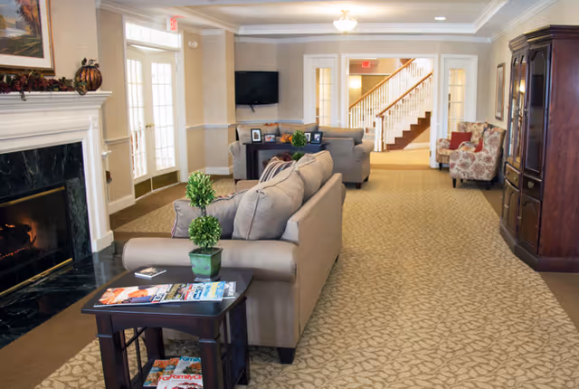 A cozy living room area in a senior living facility with beige sofas, a small table with magazines and a potted plant, a fireplace on the left, a wall-mounted TV, and a staircase in the background.
