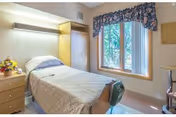 Single-occupancy nursing facility bedroom with a hospital-style bed, bedside table with flowers, window with floral valance, and built-in cabinetry.