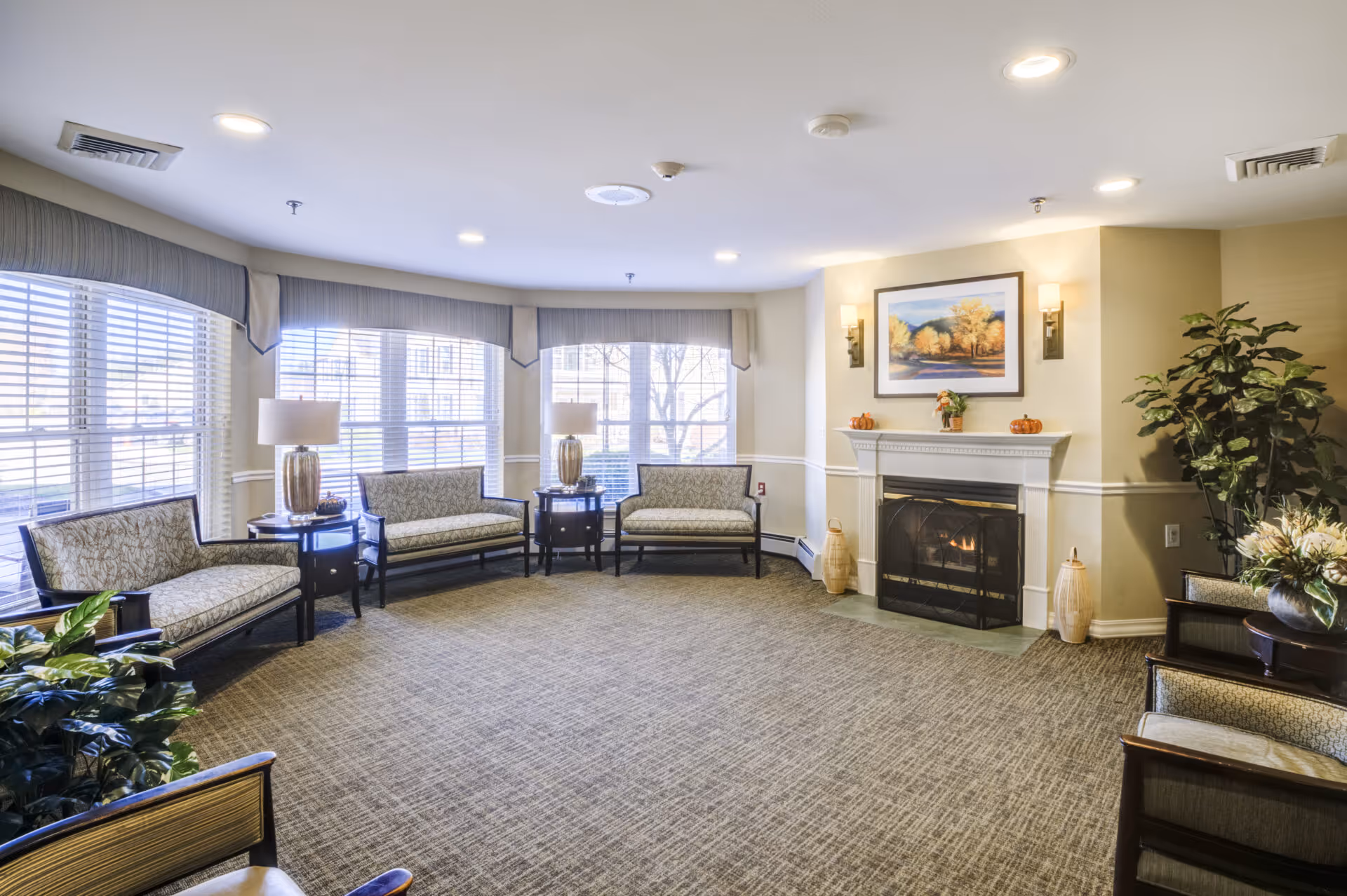 A bright and spacious senior living common area with large windows letting in natural light. The room features several cushioned chairs and loveseats arranged around the perimeter, two table lamps on side tables, a fireplace with a painting of autumn trees above it, and decorative plants in the corners.