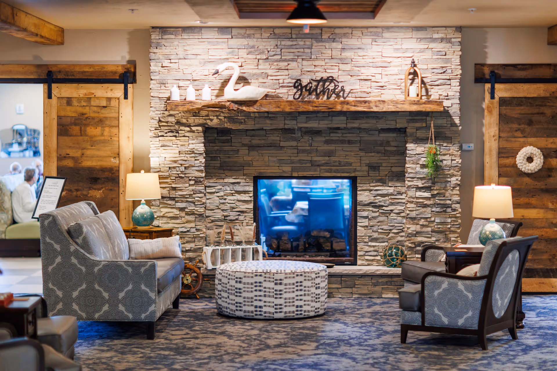 Cozy senior living facility common area with a stone fireplace, a wooden mantel decorated with a white swan figurine, candles, and a 'gather' sign. The room features patterned upholstered chairs and a sofa, a round patterned ottoman, two table lamps with blue bases, rustic wooden sliding barn doors, and a carpeted floor.