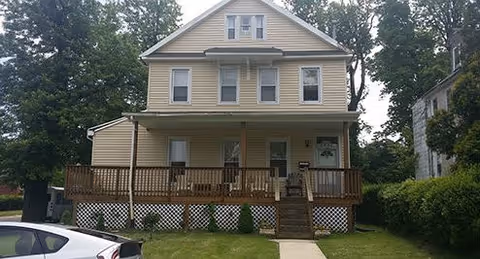 A three-story beige house with a wooden front porch and steps leading up to the entrance. The house is surrounded by trees and has a small lawn in front with a sidewalk leading to the porch. A white car is partially visible on the left side of the image.