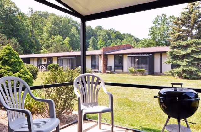 View from a covered patio with two white plastic chairs and a black charcoal grill, overlooking a grassy yard with bushes and trees, and a single-story building in the background under a clear sky.