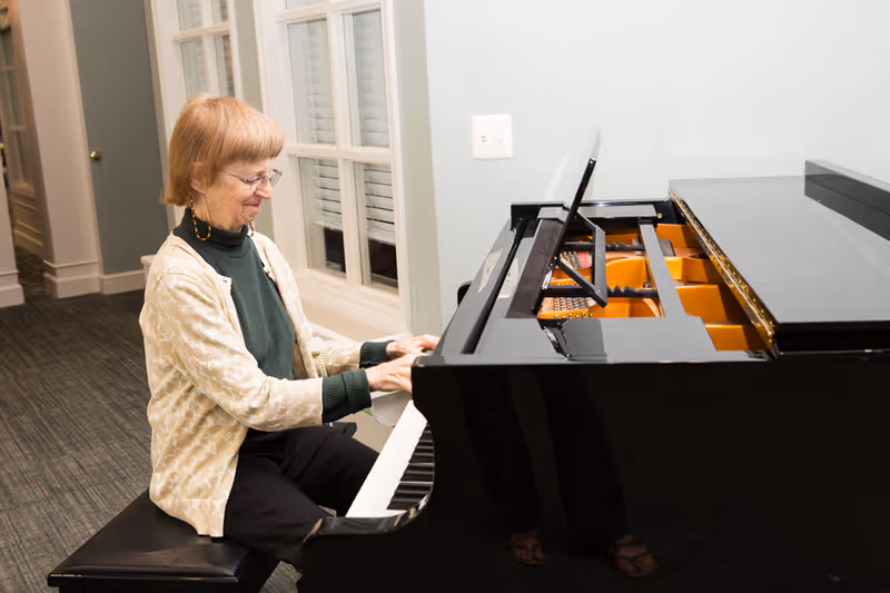 An elderly woman with glasses and short light brown hair is sitting on a black piano bench playing a grand piano in a well-lit room with carpeted floor and white walls. There is a window with white blinds behind her.