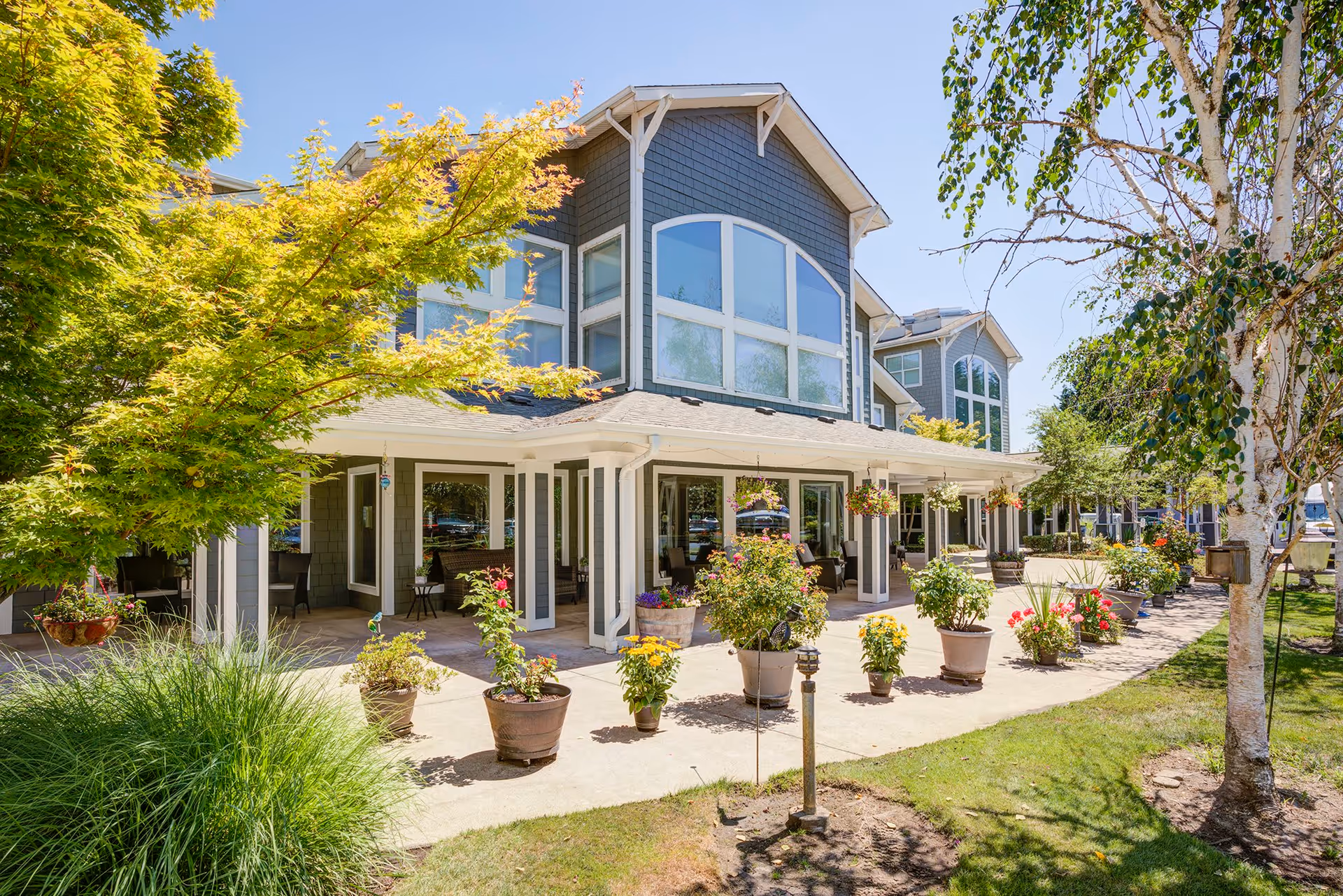 Exterior view of a senior living facility building with large windows and a covered patio area. The patio is decorated with numerous potted plants and hanging flower baskets. Trees and green grass surround the walkway leading to the entrance under a clear blue sky.