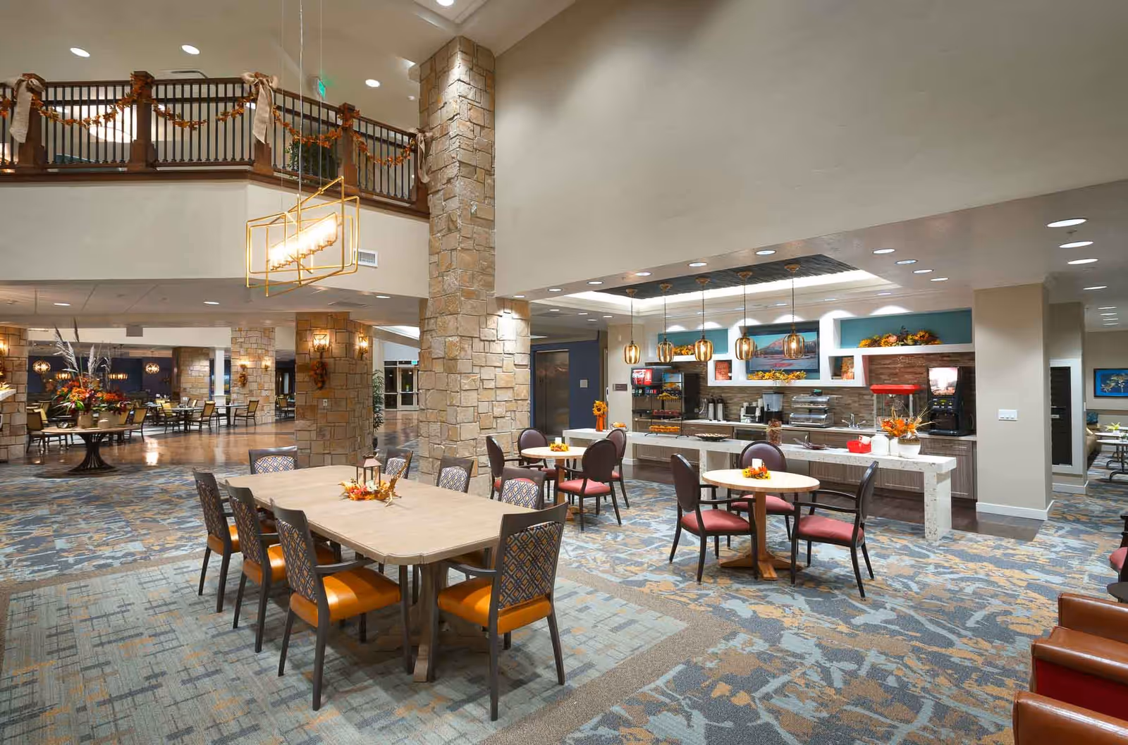 A spacious and well-lit dining area in a senior living facility with multiple tables and chairs arranged for seating. The room features stone pillars, a patterned carpet, and a serving counter with pendant lights above it. The decor includes autumn-themed floral arrangements and garlands, and there is a second-floor balcony with railings visible.
