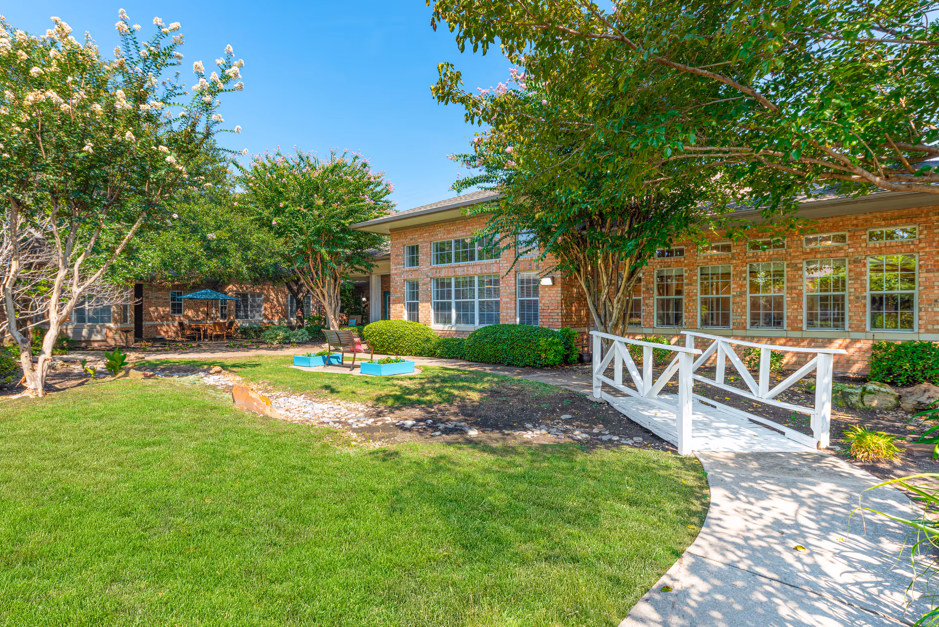 A sunny outdoor garden area at Spring Creek Assisted Living featuring a well-maintained lawn, several trees, a small white wooden bridge over a dry creek bed, and a brick building with large windows in the background. There are also a few chairs and a table under an umbrella visible in the shaded area.