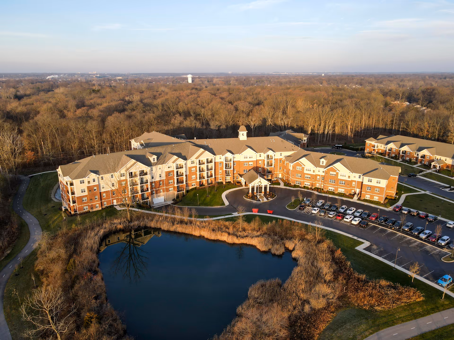 Aerial view of Inniswood Village senior living facility showing a large, multi-story building with a clock tower and covered entrance. The building is surrounded by parking lots with cars, a pond with surrounding vegetation, and a wooded area in the background under a clear sky.