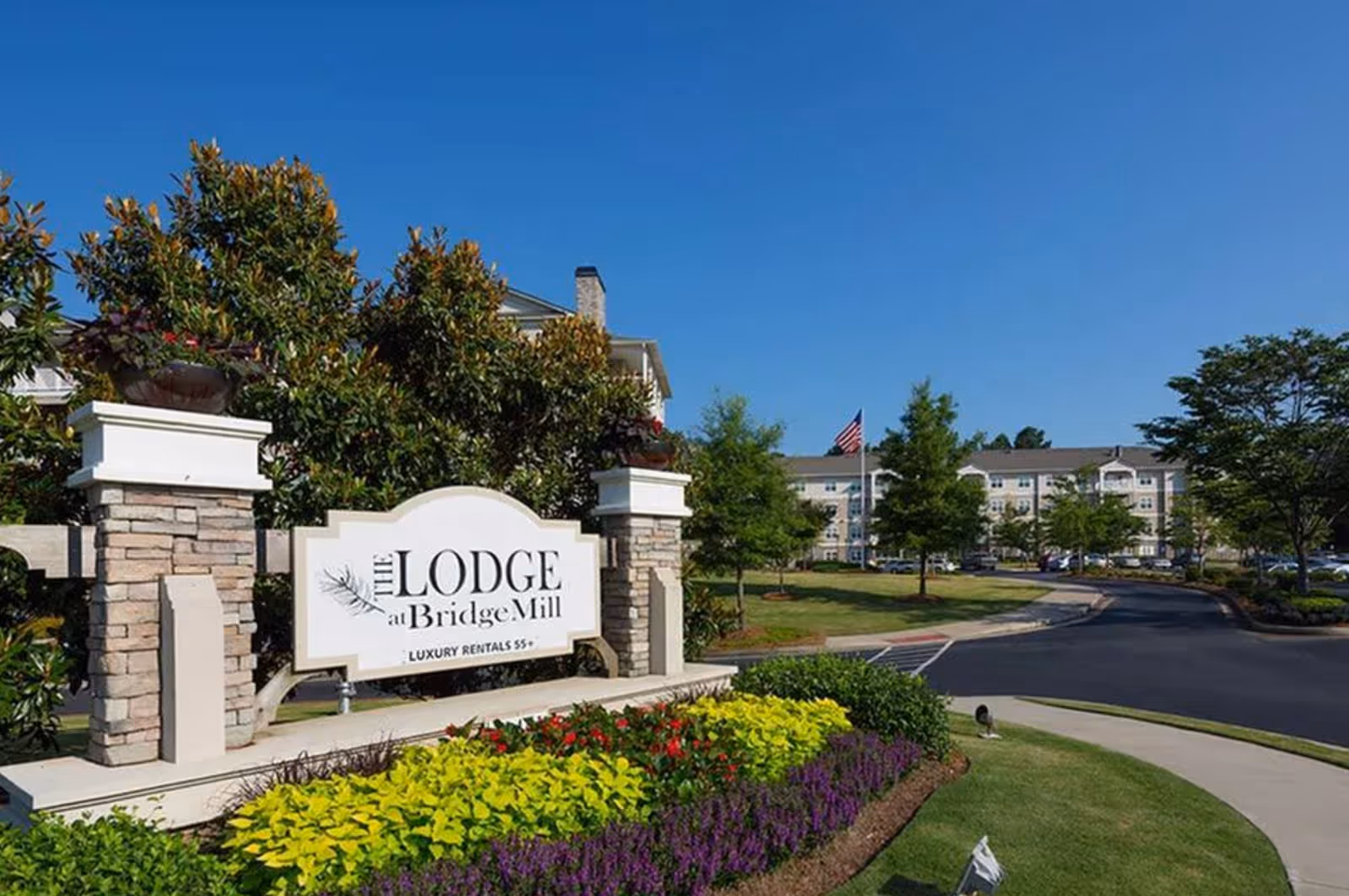 Entrance sign for The Lodge at BridgeMill luxury rentals 55+ surrounded by landscaped flowers and greenery, with a large building and American flag visible in the background under a clear blue sky.