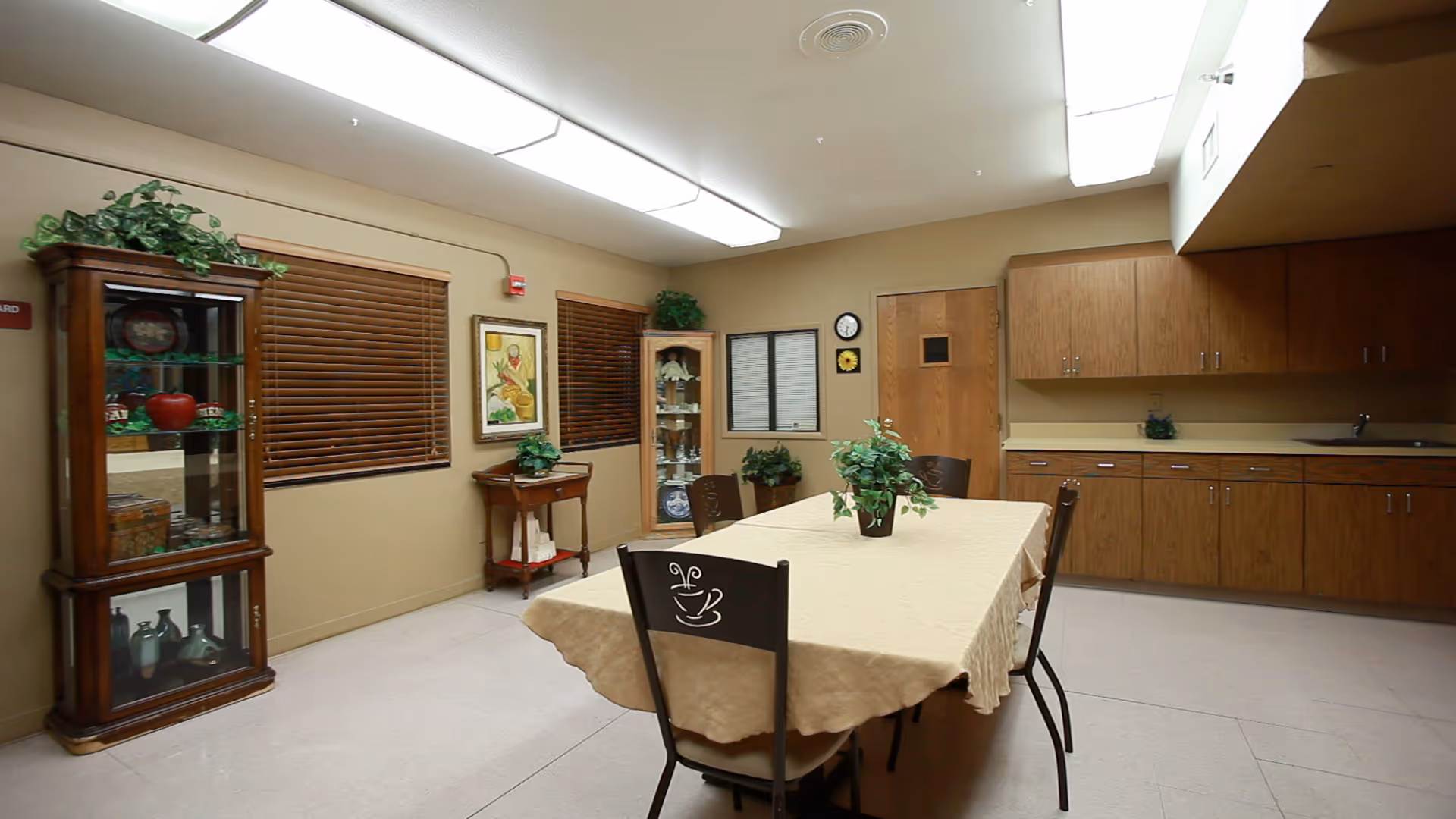 A well-lit room with a rectangular table covered with a beige tablecloth and four chairs around it. The room has wooden cabinets along one wall with a sink, two glass display cabinets with decorative items, two windows with wooden blinds, a small side table with a plant, and a door. The ceiling has fluorescent lighting panels.