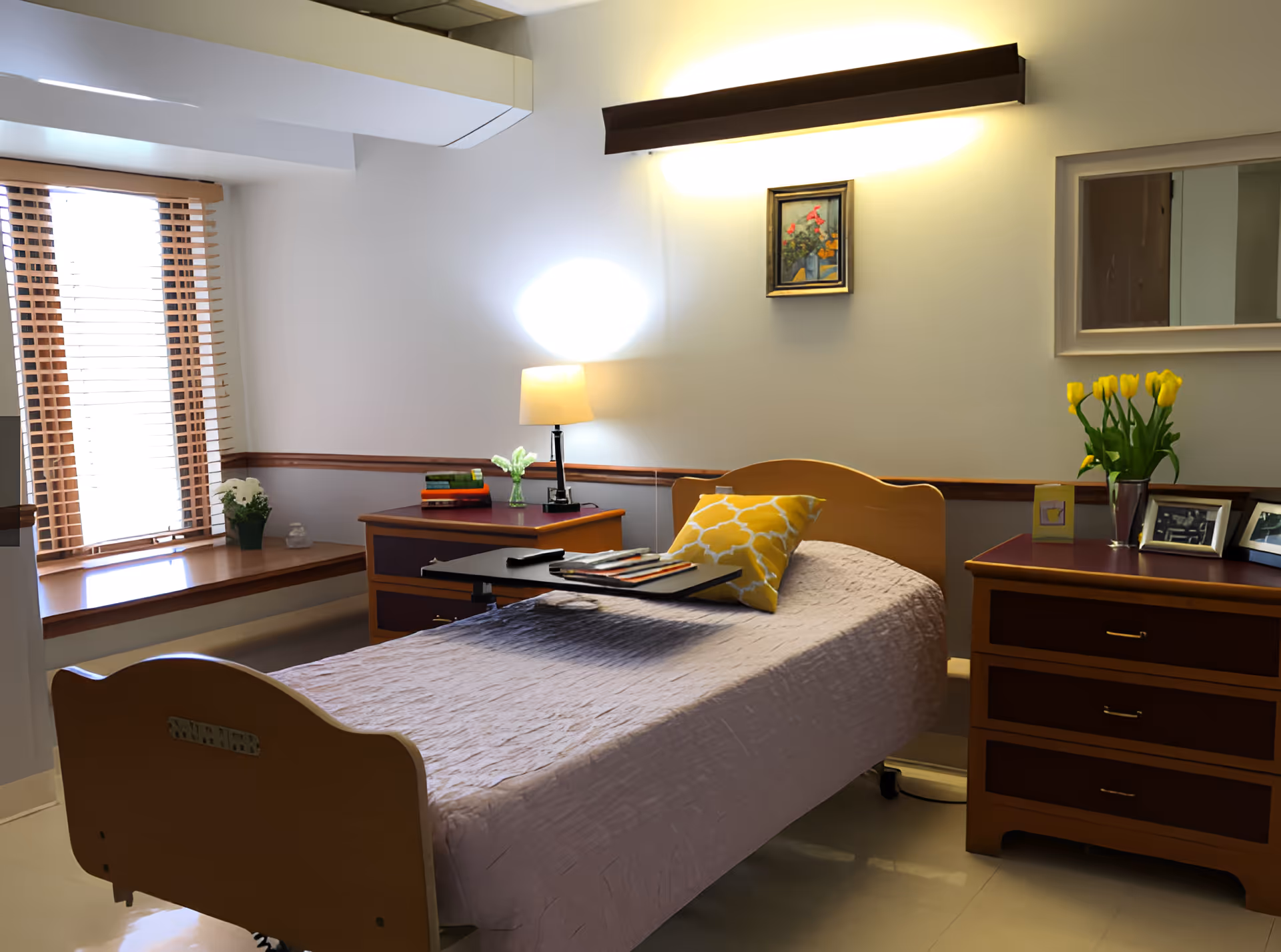 A neatly arranged bedroom in a senior living facility with a single bed covered in a light purple quilt and a yellow patterned pillow. There is a wooden bedside table with a lamp, books, and a small vase with flowers. Another wooden dresser with yellow tulips in a vase and framed photos is on the right side. A window with wooden blinds allows natural light into the room. A small painting and a mirror hang on the wall above the bed.