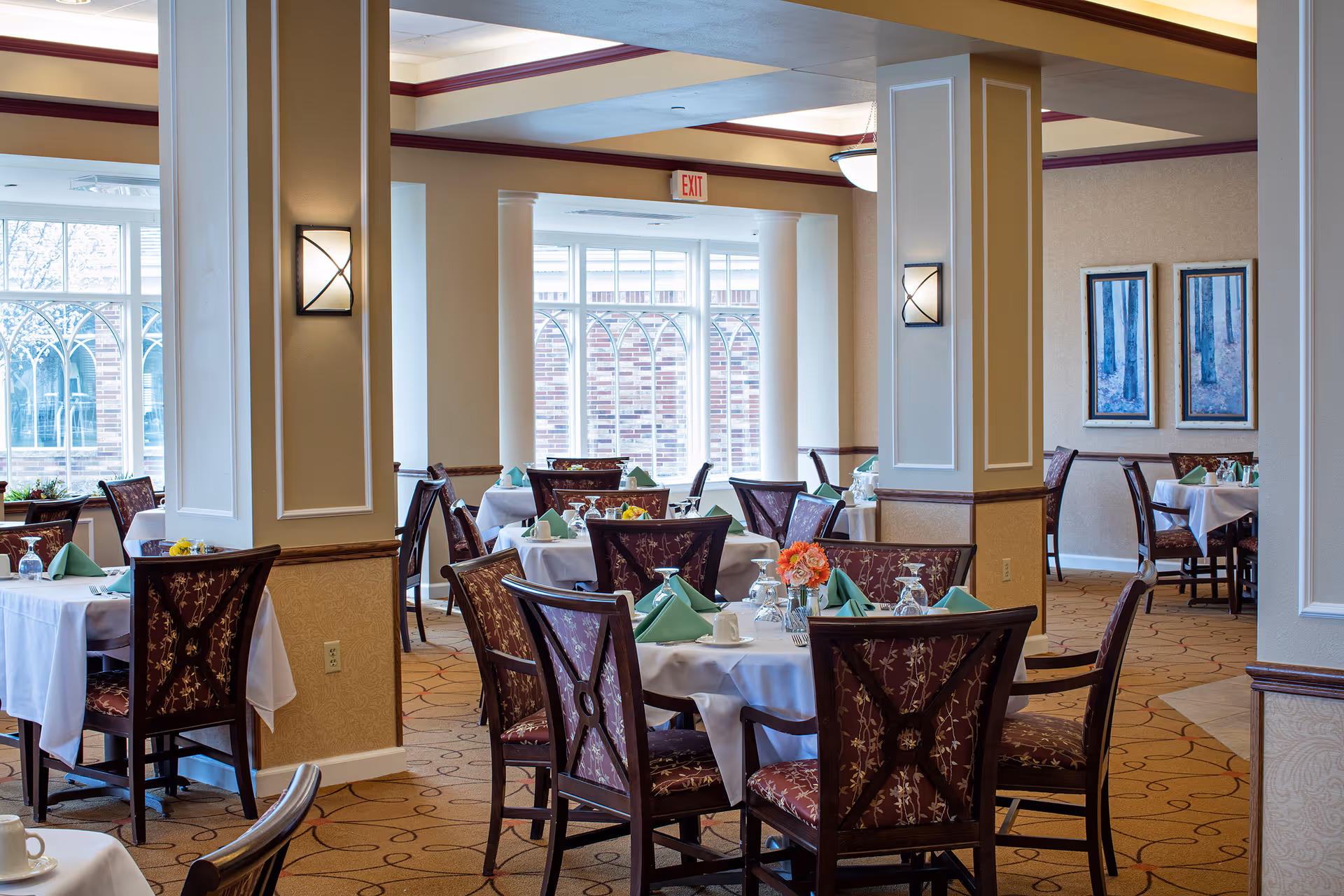 A bright dining room with multiple round tables covered in white tablecloths, each set with green folded napkins, glassware, and floral centerpieces. The room features large windows with decorative arches, beige walls with framed artwork, and wooden chairs with patterned upholstery.