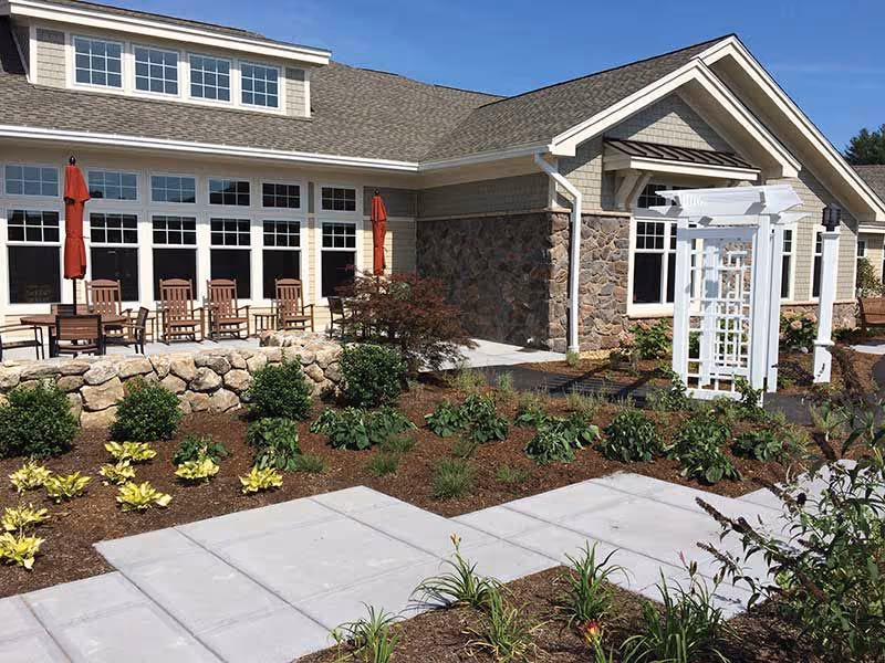 Stone-and-siding building front with a patio of chairs and umbrellas, white pergola, and landscaped walkway.