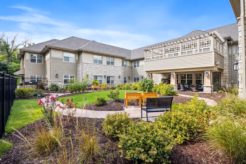 Outdoor garden area at The Grande at Creve Coeur featuring a well-maintained lawn, various shrubs and flowers, a paved walking path, raised garden beds, and a black metal bench. The background shows a two-story building with beige siding and stone accents under a clear blue sky.