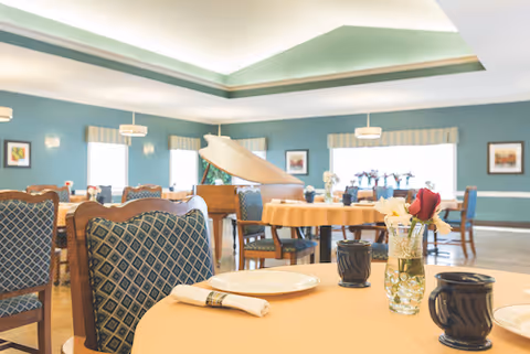 Bright dining room with round tables set for meals, upholstered chairs, and a grand piano in the background.