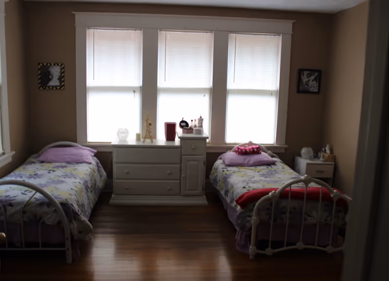A bedroom with two twin beds, a white dresser beneath three windows, and hardwood floors.