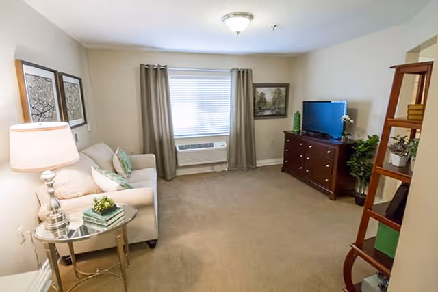 A cozy living room with beige carpet and walls, featuring a white sofa with green and white pillows, a round glass side table with a lamp and books, a window with gray curtains, a wooden dresser with a flat-screen TV on top, and a wooden ladder shelf with decorative items.