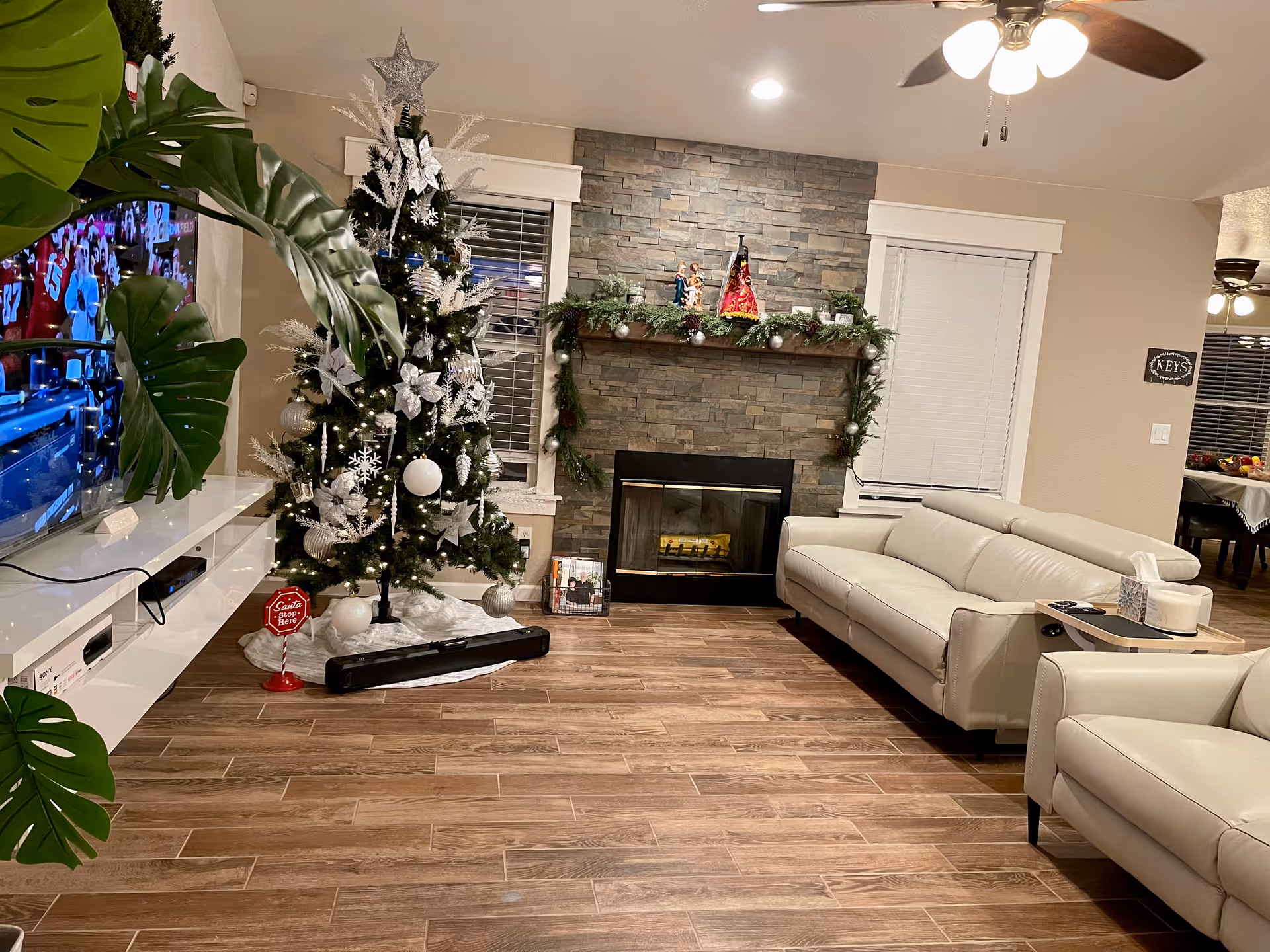 A cozy living room decorated for Christmas with a tall Christmas tree adorned with white and silver ornaments and a star on top. There is a stone fireplace with a wooden mantel decorated with garland and Christmas figurines. The room has light-colored leather sofas, a ceiling fan with lights, and a TV mounted on the wall showing a sports game. The floor is wooden and there are large green leaves partially visible on the left side.