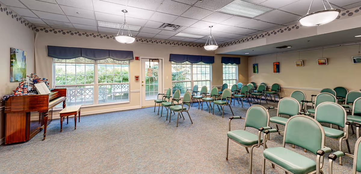 A spacious activity room with rows of green chairs, large windows, and a piano near the entrance.