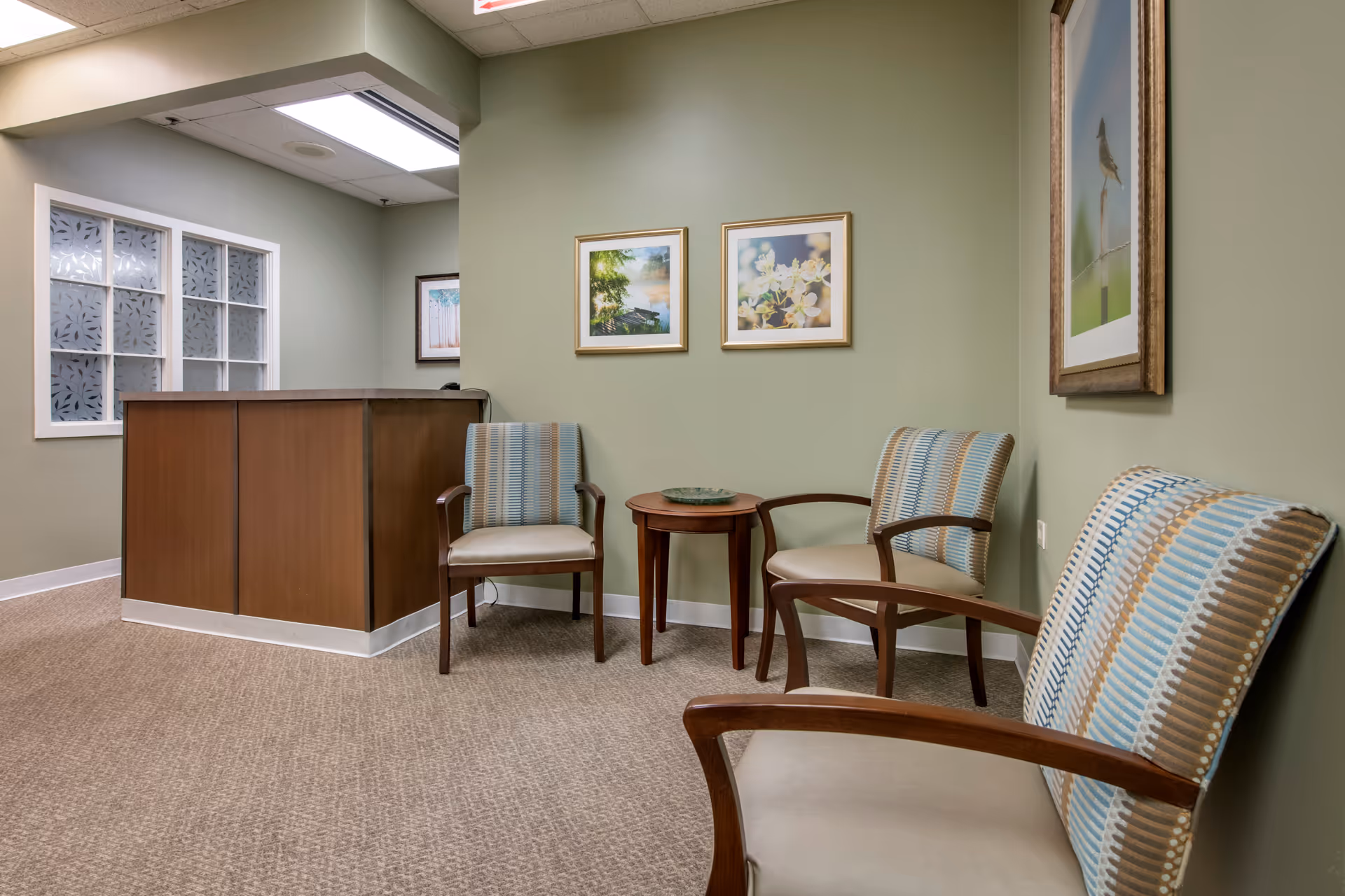 A waiting area with three cushioned chairs featuring striped upholstery in blue, beige, and brown tones. There is a small wooden round table with a decorative bowl between two of the chairs. The walls are painted light green and decorated with framed pictures of nature scenes. A wooden reception desk is visible in the background under a ceiling light.