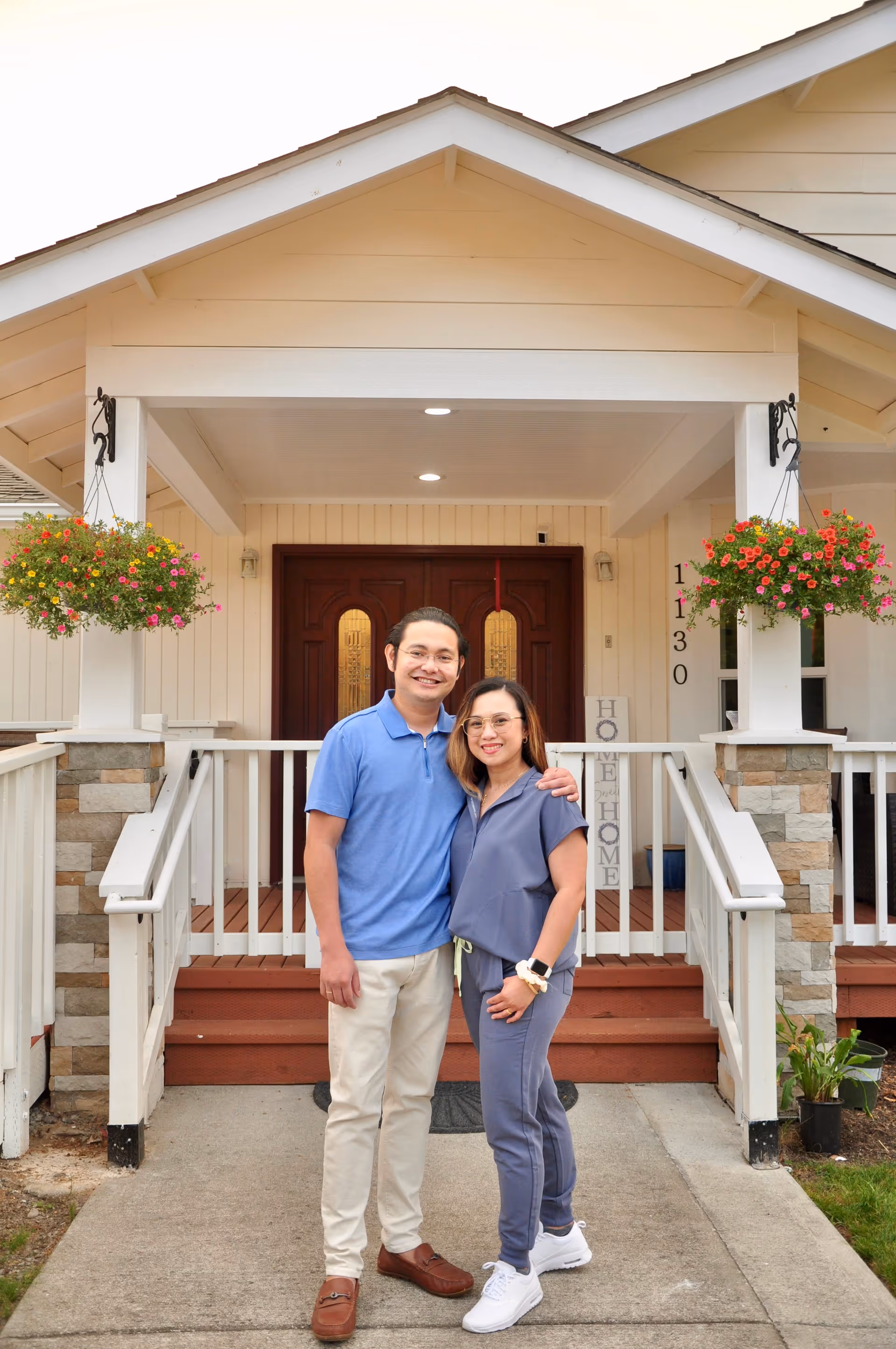 A man and a woman standing arm in arm, smiling in front of the entrance to a house with a covered porch. The porch has white railings, stone pillars, and hanging flower baskets with colorful flowers. The house has beige siding and a double wooden front door with decorative glass panels.