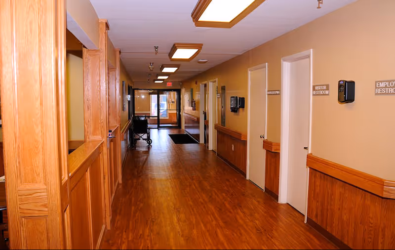 A well-lit hallway in a senior living facility with wooden flooring and wood paneling on the lower half of the walls. Several doors line the right side of the hallway, including ones labeled 'Visitor Restroom' and 'Employee Restroom'. The hallway leads to a glass exit door at the far end.