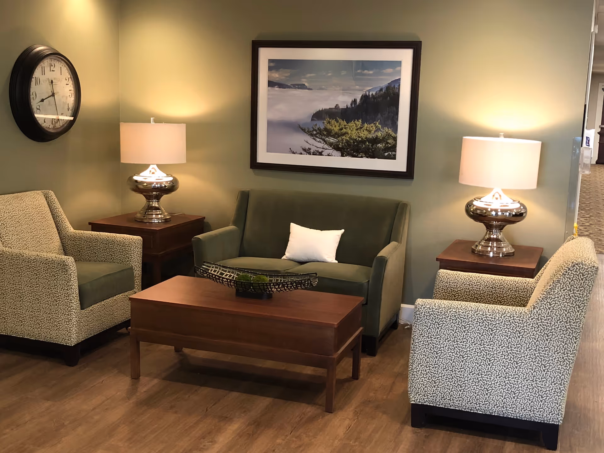 A cozy sitting area with a green loveseat, two patterned armchairs, a wooden coffee table, and two side tables with silver lamps. A large clock hangs on the left wall and a framed landscape photograph is centered above the loveseat. The walls are painted light green and the floor is wood.