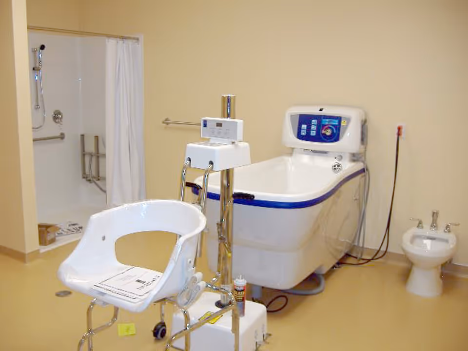 A clinical bathroom with a medical bathing chair, a specialized bathtub with control panel, a bidet, and a shower area with grab bars and a curtain.