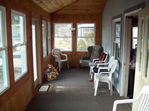 A sunroom or enclosed porch with wood-paneled walls and ceiling, featuring several white plastic chairs and one cushioned armchair with a plaid pattern. There is a floor lamp in the corner and a basket of flowers on the floor near the windows. The room has multiple windows allowing natural light and a view of the outdoors.
