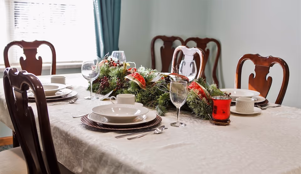 Dining table set with plates, wine glasses and a festive evergreen centerpiece surrounded by wooden chairs.