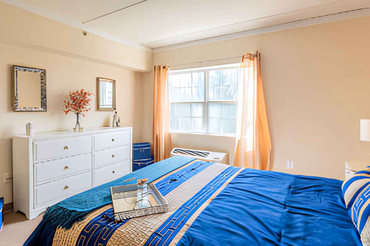 Sunlit bedroom with a large bed dressed in blue linens, a white dresser with mirrors, and a window with peach curtains.