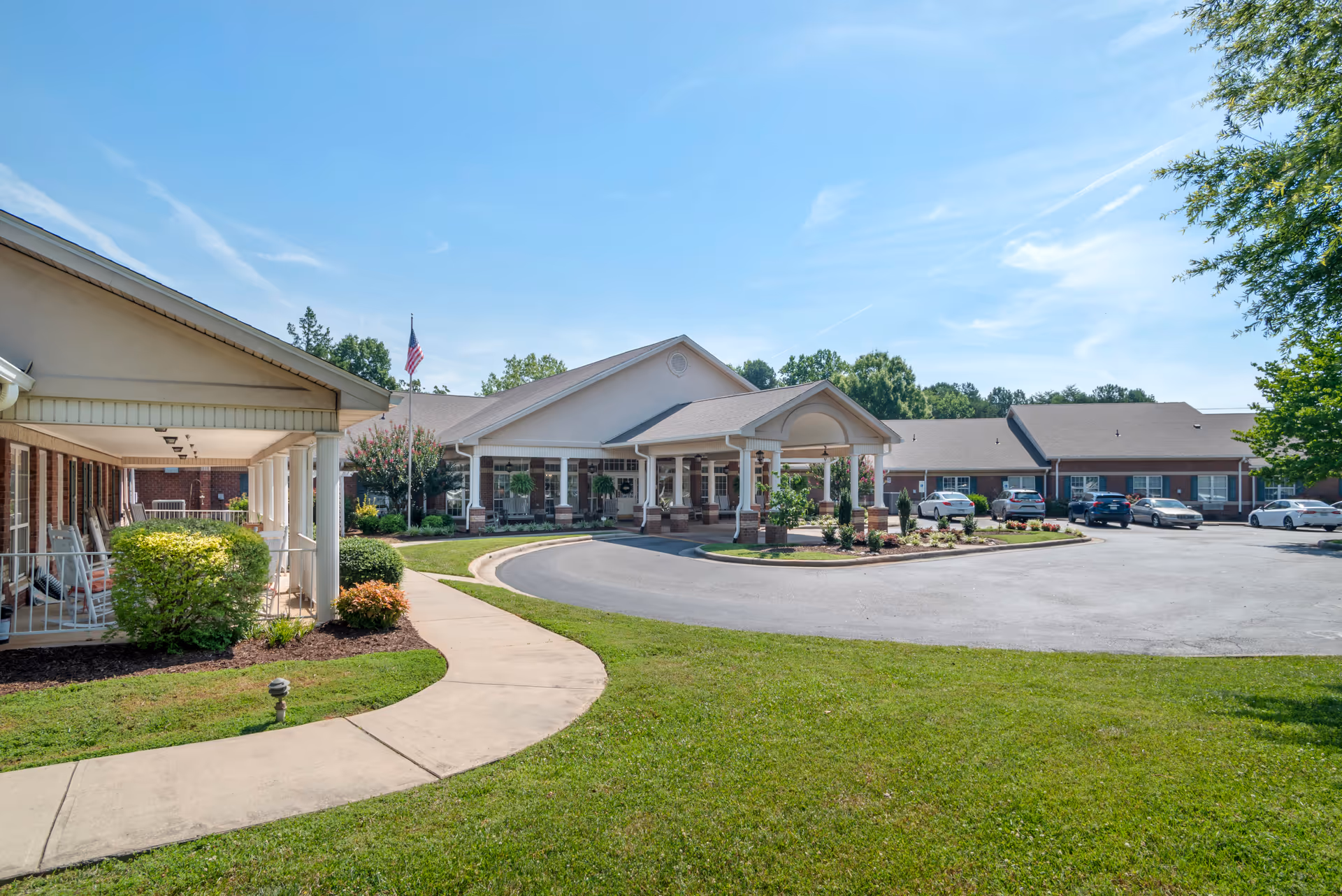 Front entrance of a senior living facility with a covered porte-cochere, circular driveway, landscaping, and parked cars.