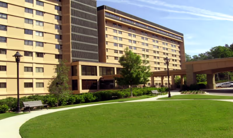 Exterior view of a multi-story beige building with many windows, surrounded by green grass, trees, benches, and lamp posts under a clear blue sky.