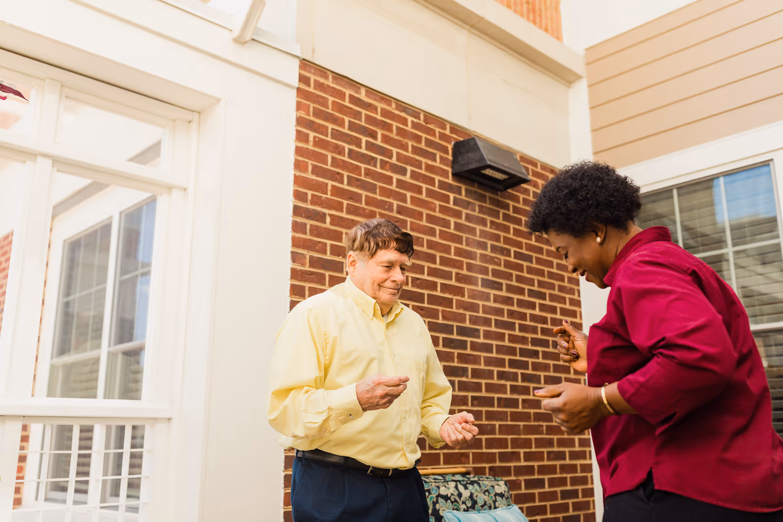 An elderly man in a yellow shirt and dark pants and a woman in a red shirt are standing and interacting happily outside a brick building with white window frames.