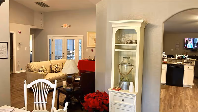 Open-plan common area with sofas and chairs, a white display cabinet, and a kitchen visible through an archway.