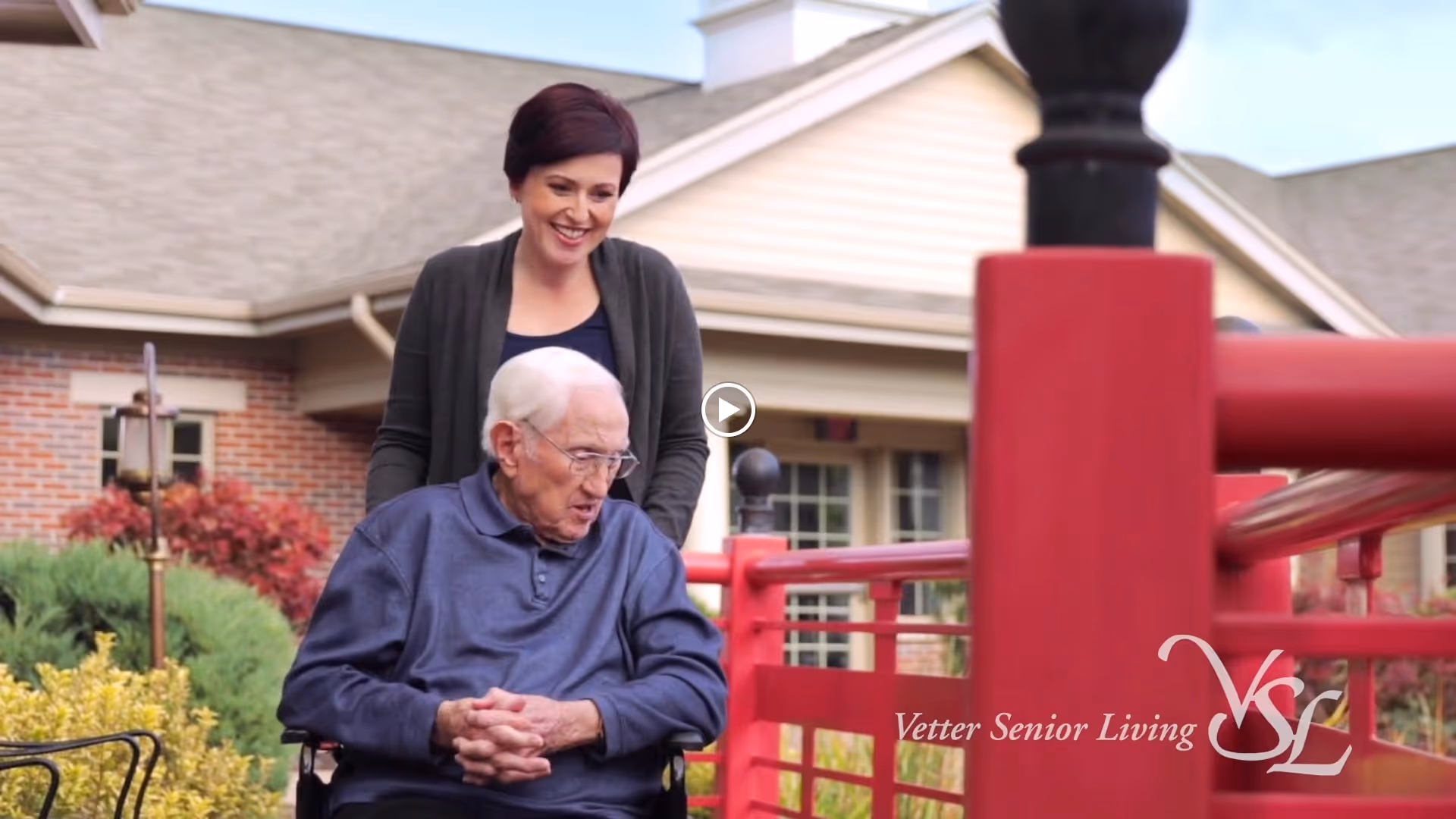 An elderly man in a wheelchair is outdoors, accompanied by a smiling woman standing behind him. They are near a red railing with a building and greenery in the background.