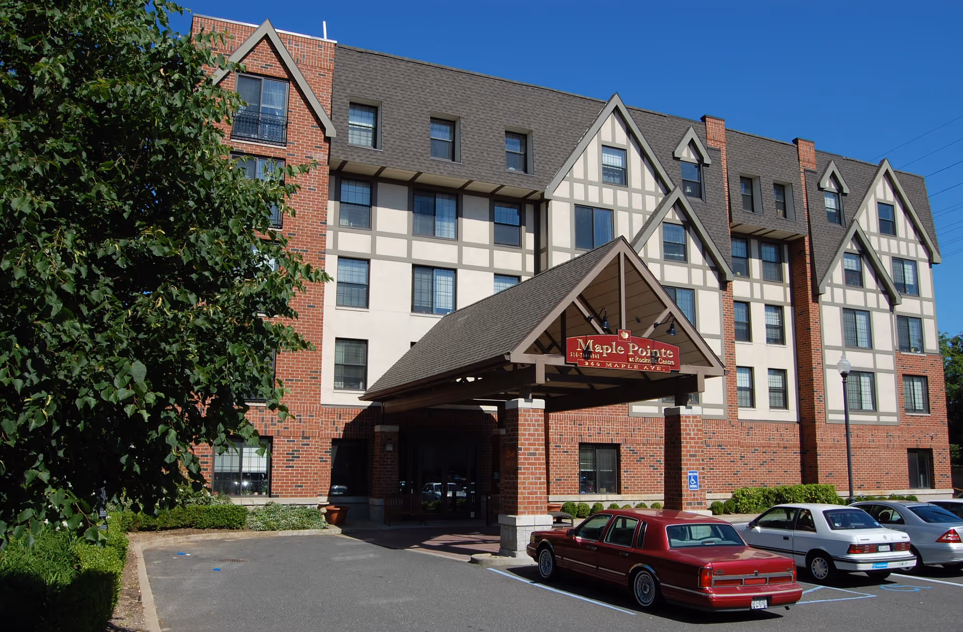 Exterior front view of Maple Pointe Senior Living facility, a multi-story building with a brick and beige facade, a covered entrance with a sign displaying the facility name, and several parked cars in front under a clear blue sky.
