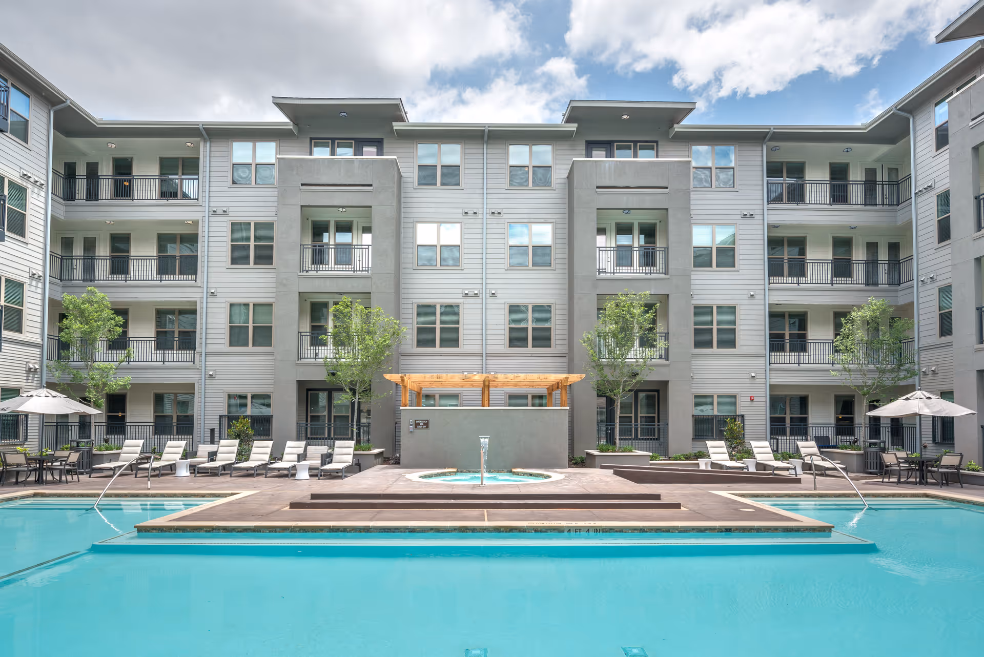 Outdoor swimming pool area with lounge chairs and umbrellas, surrounded by a multi-story residential building with balconies and windows under a partly cloudy sky.