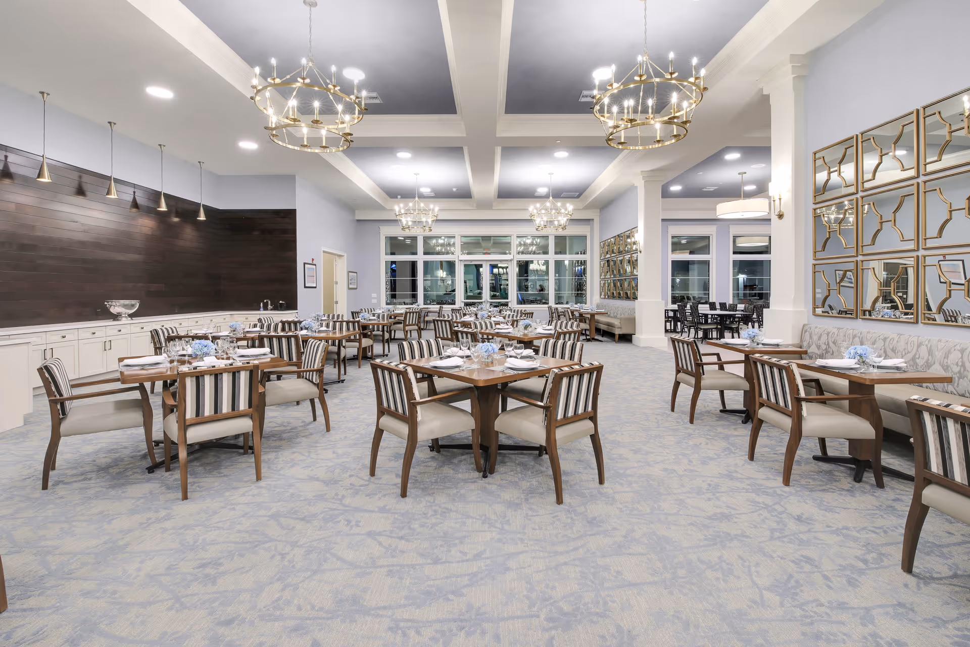 A spacious and elegant dining room in a senior living facility with multiple wooden tables and striped cushioned chairs arranged neatly. The room features soft blue carpeting, large windows, decorative mirrors on the walls, and modern chandeliers hanging from the ceiling. The tables are set with white napkins, plates, and small floral centerpieces.