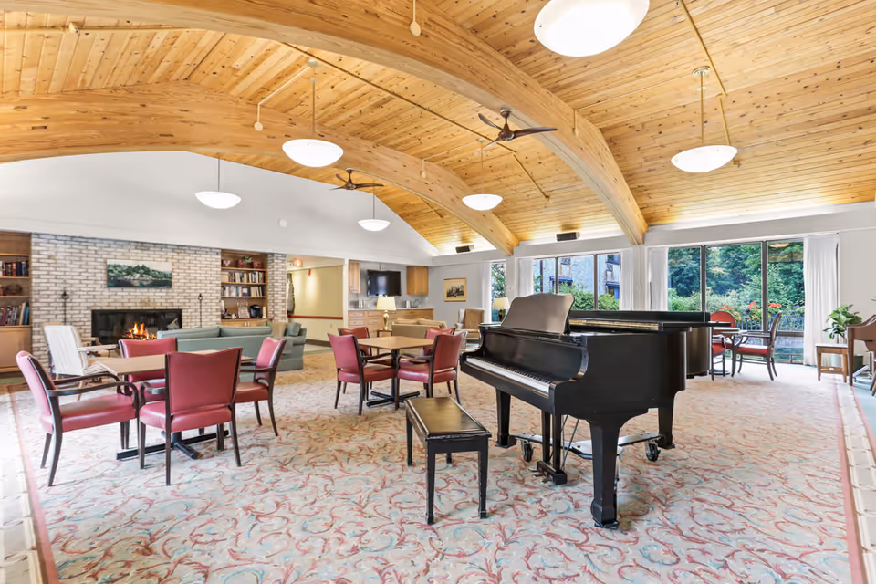 Spacious common area with a wooden vaulted ceiling, a black grand piano with a bench, several tables with red chairs, a fireplace with a brick surround, bookshelves, and large windows showing greenery outside.
