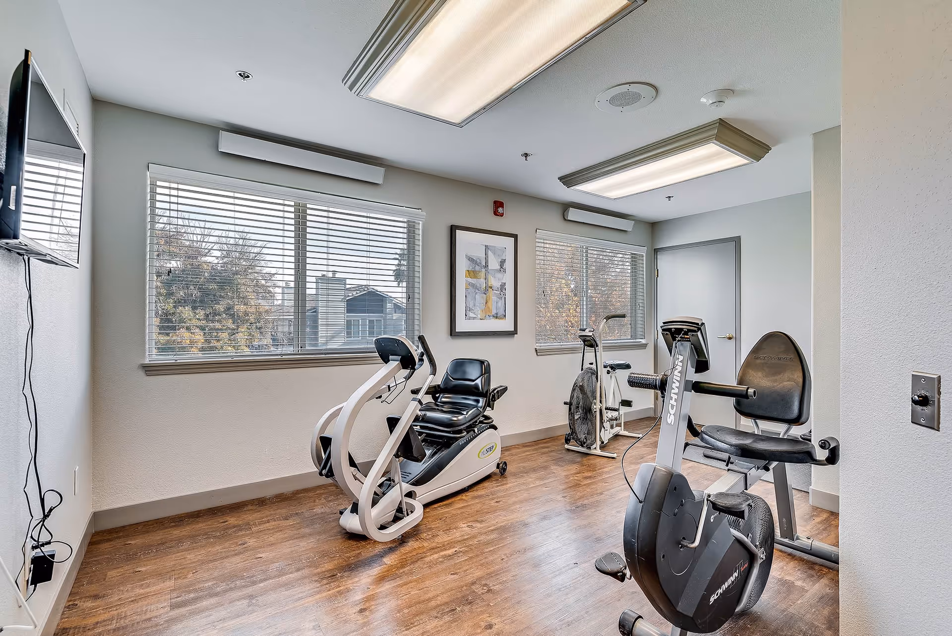 A small fitness room with exercise equipment including a recumbent bike, an upright stationary bike, and an air resistance exercise bike. The room has wood flooring, two large windows with blinds, a wall-mounted TV, and fluorescent ceiling lights. There is a framed abstract painting on the wall between the windows.