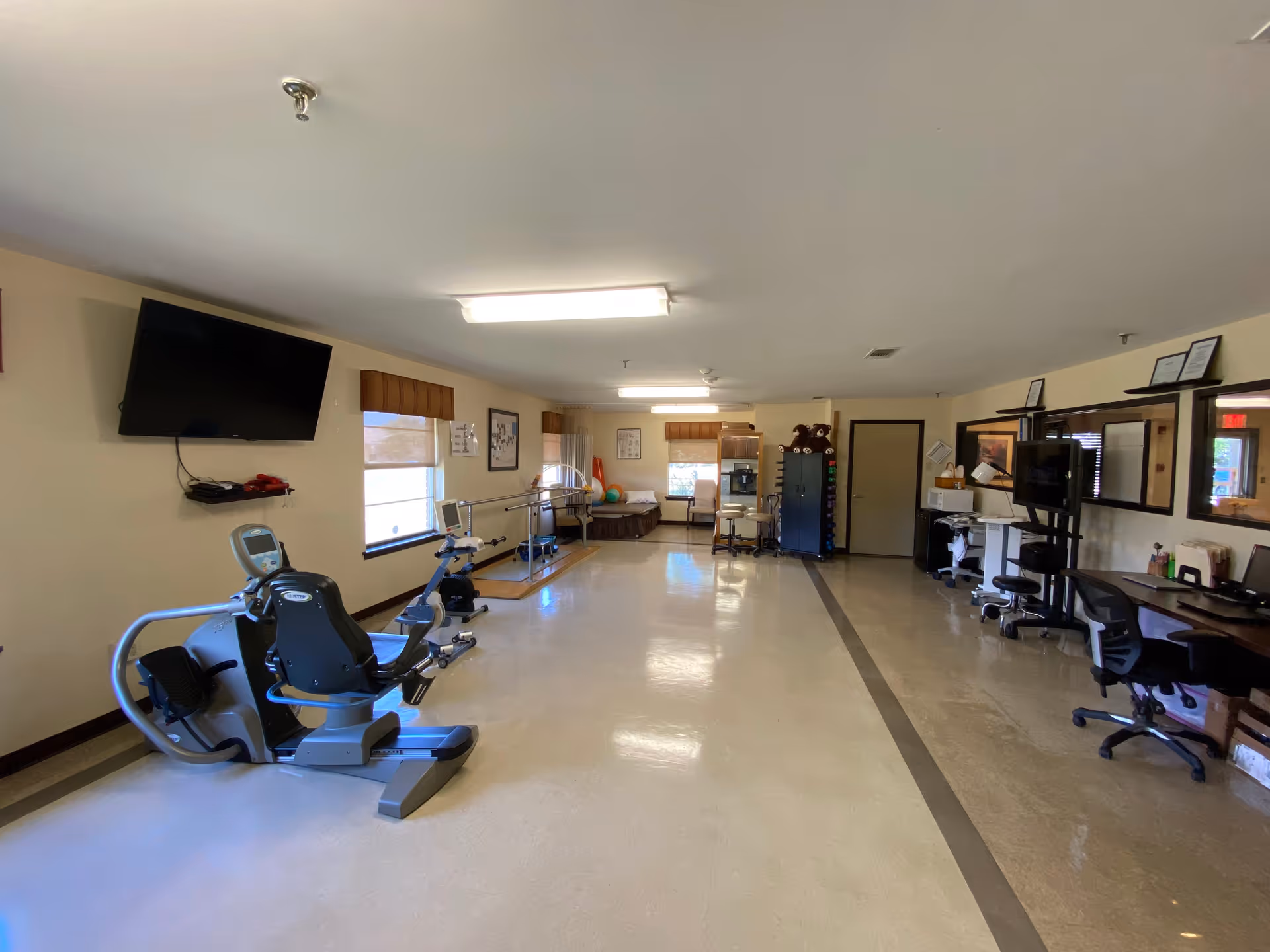 Spacious interior therapy and exercise room with a recumbent bike, therapy equipment, desks and a wall-mounted TV.