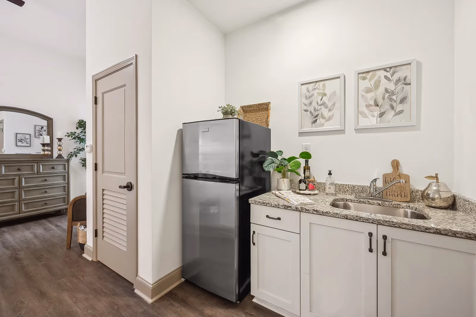 Compact kitchenette featuring a stainless steel refrigerator, granite countertop with sink and cabinets, decorative plants and framed botanical prints, with a dresser visible in the adjoining room.