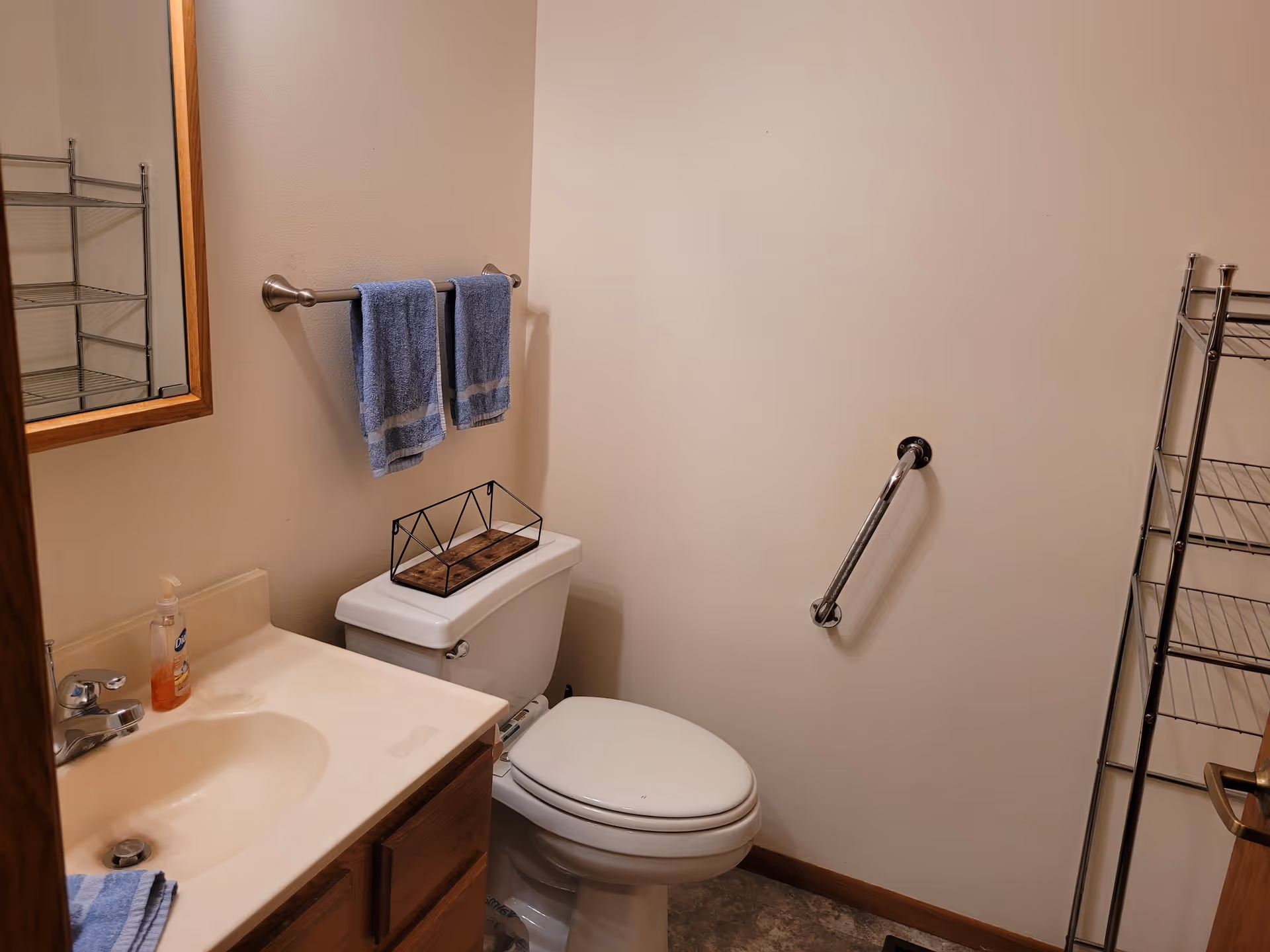 Small bathroom with a white toilet, a sink with a beige countertop, a mirror with a wooden frame, two blue hand towels hanging on a metal towel rack, a metal grab bar on the wall, and a metal shelving unit.
