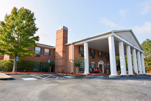 Exterior view of a two-story brick building with large white columns at the entrance, a chimney, and a tree in the parking lot with handicap parking spaces visible.