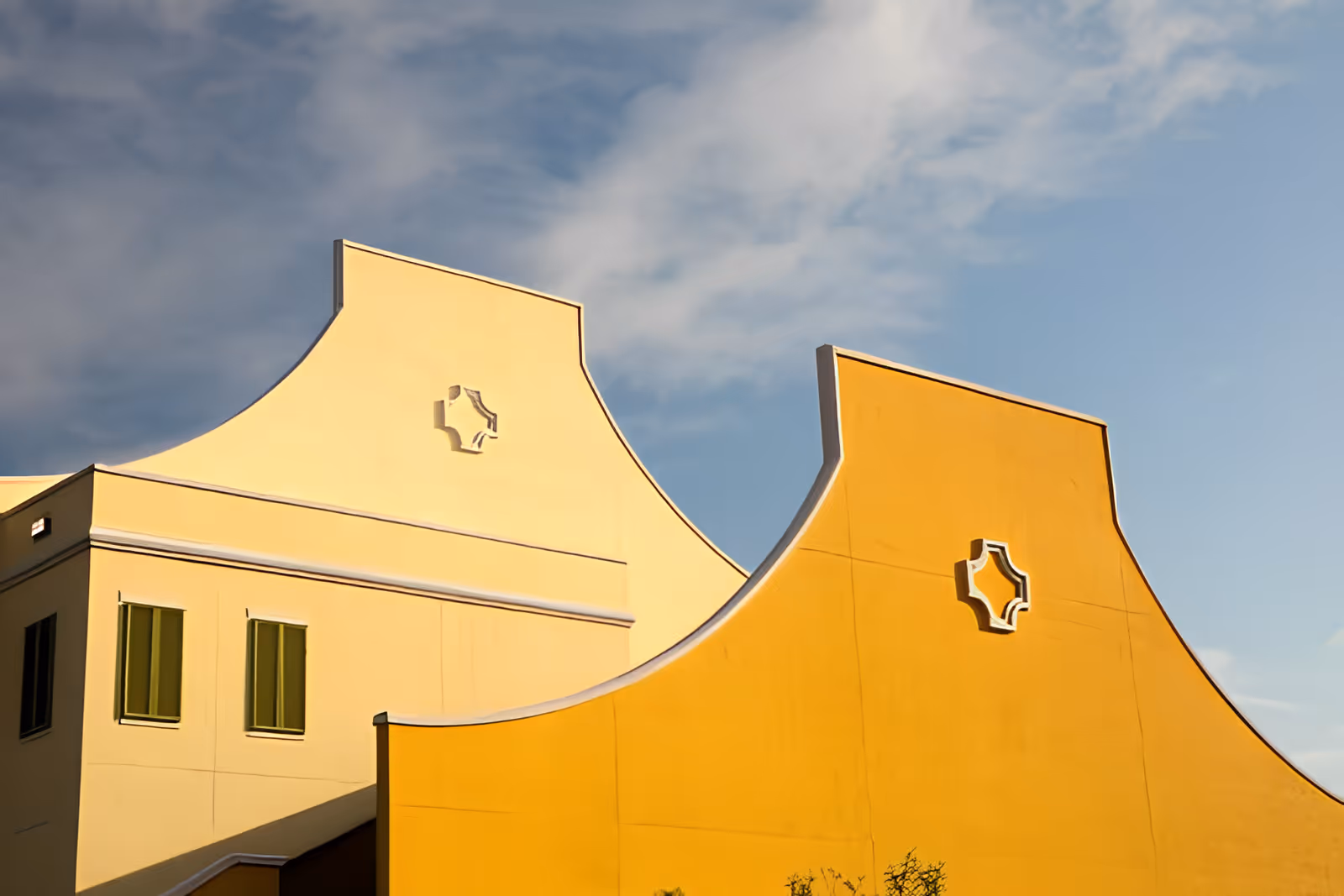 Exterior view of a building with two distinct yellow and orange walls featuring curved tops and decorative cut-out shapes near the top, under a partly cloudy sky.