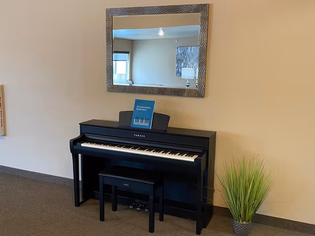 A black Yamaha digital piano with a matching bench placed against a beige wall. Above the piano is a large rectangular mirror with a textured frame reflecting part of the room, including a window and a lamp. To the right of the piano is a potted green plant with tall grass-like leaves.