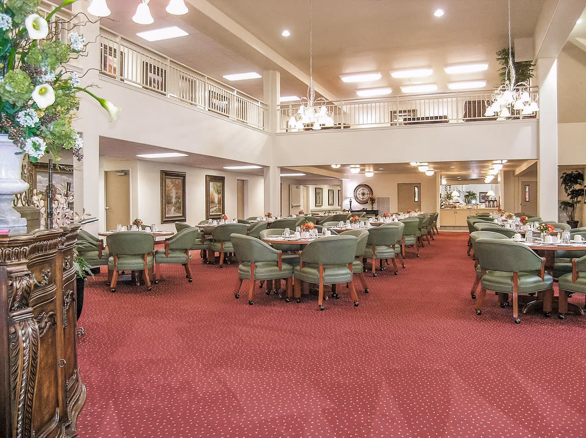 Spacious dining room with multiple round tables set with cups, plates, and small flower arrangements. The room has green cushioned chairs, red carpet flooring, chandeliers hanging from the ceiling, and a balcony railing overlooking the area. There are paintings on the walls and a large floral arrangement on a wooden cabinet to the left.